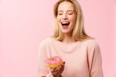 Pembe arka planda izole edilmiş donutla poz verirken gülen neşeli sarışın kız.