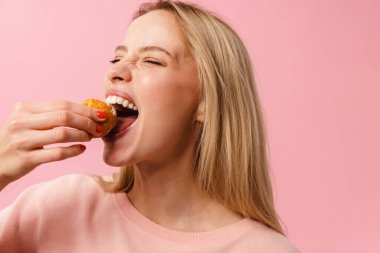 Mutlu sarışın kız pembe arka planda izole bir şekilde kamerada donut yiyor.