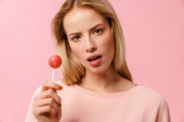 Pembe arka planda izole edilmiş lolipopla poz veren kafası karışmış sarışın kız.