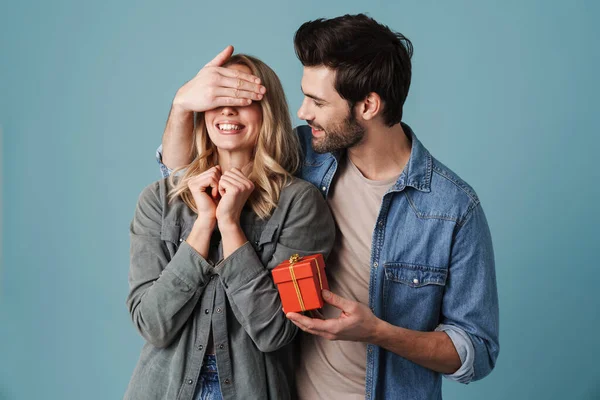 Young happy man with gift box covering eyes of excited girl isolated over blue background