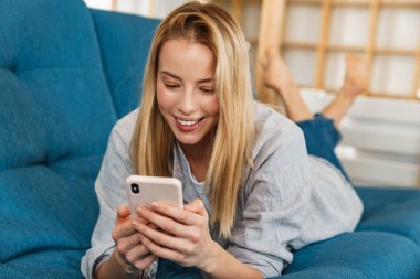 Evdeki kanepede yatarken gülümseyen sarışın kadın cep telefonu kullanıyor.