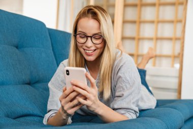 Evdeki kanepede yatarken gülümseyen sarışın kadın cep telefonu kullanıyor.