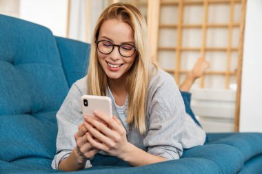 Evdeki kanepede yatarken gülümseyen sarışın kadın cep telefonu kullanıyor.