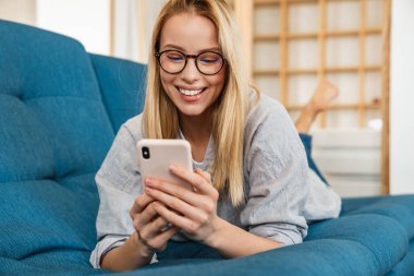 Evdeki kanepede yatarken gülümseyen sarışın kadın cep telefonu kullanıyor.