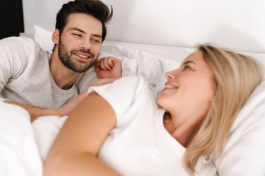 Mutlu genç çift evde yatakta yatarken gülümseyip birbirlerine bakıyorlar.
