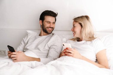 Mutlu genç çift evde yatarken gülümsüyor ve cep telefonu kullanıyor.