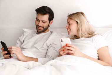Mutlu genç çift evde yatarken gülümsüyor ve cep telefonu kullanıyor.