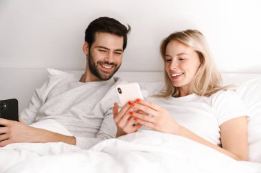 Mutlu genç çift evde yatarken gülümsüyor ve cep telefonu kullanıyor.