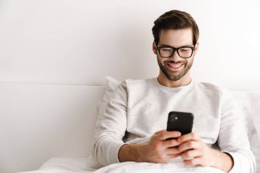 Gözlüklü gülümseyen genç adam evde dinlenirken cep telefonu kullanıyor.