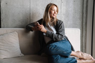 Evdeki koltukta otururken gülümseyen sarışın kadın cep telefonu kullanıyor.