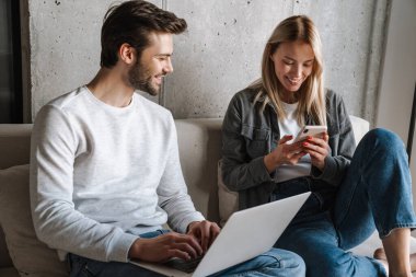 Mutlu genç çift evdeki koltukta otururken cep telefonu ve laptop kullanıyor.