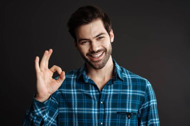 Neşeli, yakışıklı, tıraşsız, gülümseyen ve siyah arka planda iyi bir işaret gösteren adam.