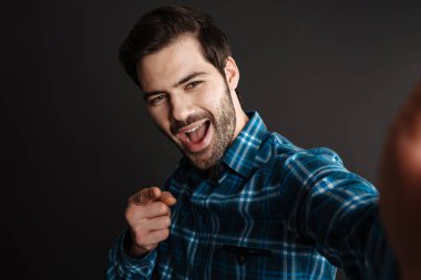 Siyah arka planda izole edilmiş selfie çekerken kamerayı işaret eden neşeli yakışıklı adam.