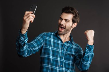 Heyecanlı adam siyah arka plandan izole edilmiş cep telefonuyla selfie çekerken kazanan hareketi yapıyor.