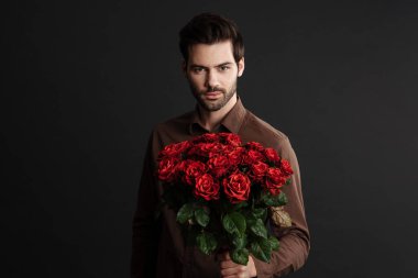 Siyah arka planda saklanmış bir buket gülle poz veren kendine güvenen yakışıklı, tıraşsız adam.