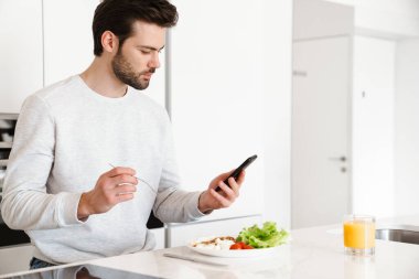 Ev mutfağında kahvaltı yaparken cep telefonu kullanan yakışıklı bir adam.