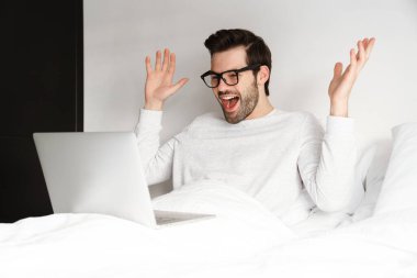 Evde dizüstü bilgisayar kullanırken el kol hareketi yapan heyecanlı genç adam.