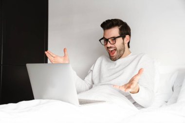 Evde dizüstü bilgisayar kullanırken el kol hareketi yapan heyecanlı genç adam.