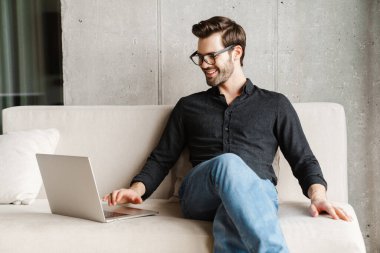 Evdeki kanepede otururken bilgisayarını kullanan traşsız bir adam.