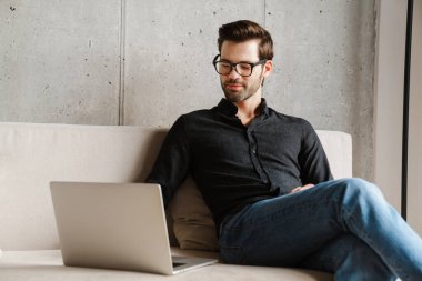 Evdeki kanepede otururken bilgisayarını kullanan traşsız, memnun bir adam.