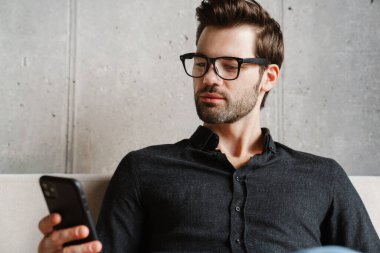 Evdeki koltukta otururken akıllı telefon kullanan ciddi, tıraşsız bir adam.