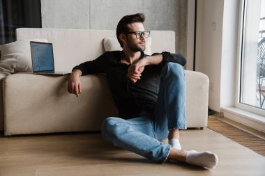 Evde yerde otururken dizüstü bilgisayar kullanan memnun bir adam.