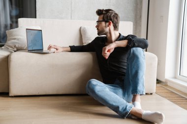 Evde yerde otururken dizüstü bilgisayar kullanan tıraşsız bir adam.