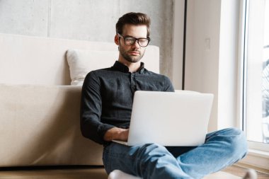 Evde otururken dizüstü bilgisayar kullanan ciddi, tıraşsız bir adam.