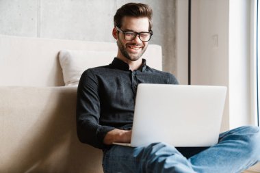 Evde yerde otururken dizüstü bilgisayar kullanan neşeli, tıraşsız adam.