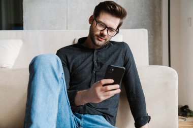 Evde otururken akıllı telefon kullanan ciddi, tıraşsız bir adam.