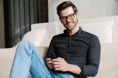 Evde yerde otururken akıllı telefon kullanan neşeli, tıraşsız adam.