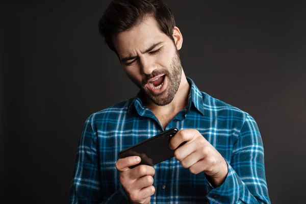 Excited handsome guy playing online game on cellphone isolated over black background