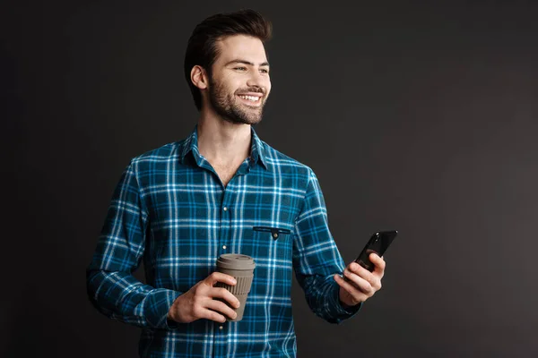 Joyful handsome guy drinking coffee takeaway and using cellphone isolated over black background