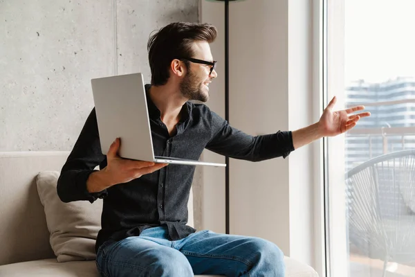 Joyful man pointing hand at window while taking video call on laptop at home