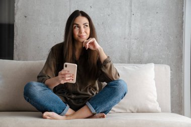 Neşeli esmer güzel kız gülümsüyor ve evdeki koltukta cep telefonu kullanıyor.