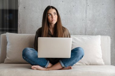 Evdeki kanepede otururken dizüstü bilgisayar kullanan esmer bir kız.