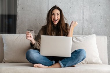 Evde dizüstü bilgisayar kullanırken göz kırpan ve kredi kartı tutan mutlu bir kız.