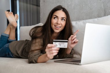 Evdeki kanepede yatarken elinde kredi kartı olan neşeli esmer kız.