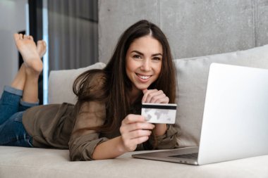 Evdeki kanepede yatarken elinde kredi kartı olan neşeli esmer kız.