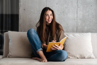 Mutlu esmer güzel kız evdeki kanepede otururken notlar yazıyor.