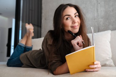 Mutlu esmer güzel kız evdeki kanepede yatarken notlar yazıyor.