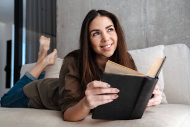 Mutlu esmer güzel kız evde kanepede uzanmış kitap okuyor.