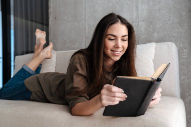 Mutlu esmer güzel kız evde kanepede uzanmış kitap okuyor.