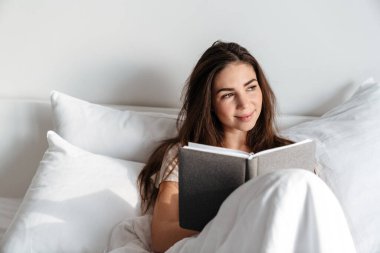 Mutlu esmer güzel kız evde yatakta otururken gülümsüyor ve kitap okuyor.