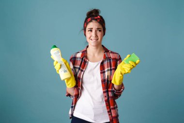 Mutsuz genç kadın eldivenli, temizlik ürünü ve süngerle mavi arka planda poz veriyor.