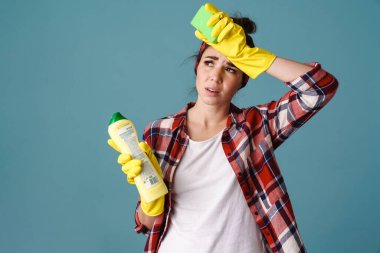 Eldivenli yorgun genç kadın mavi arka planda izole edilmiş temizlik ürünü ve süngerle poz veriyor.