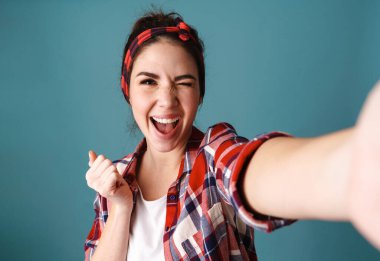 Mavi arka planda izole edilmiş selfie çekerken göz kırpan neşeli genç kadın.