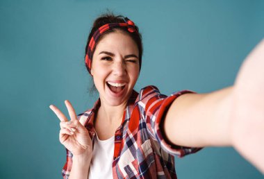 Mavi arka planda izole edilmiş selfie çekerken barış işareti gösteren neşeli genç bir kadın.