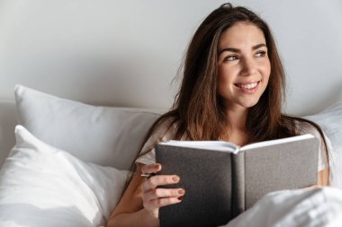 Neşeli beyaz kadın evde dinlenirken gülümsüyor ve kitap okuyor.