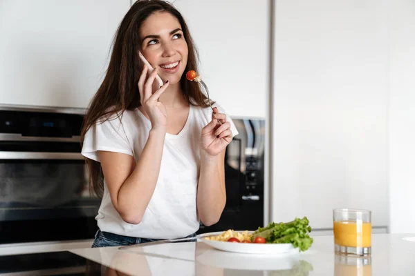 Joyful caucasian woman talking on cellphone while having lunch at home kitchen